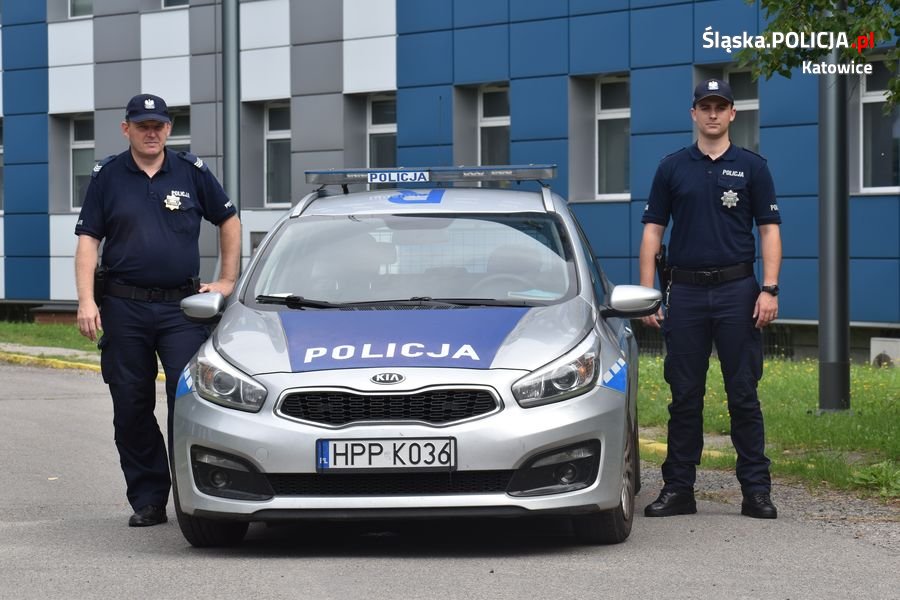 Na zdjęciu widać radiowóz po bokach radiowozu stoją policjanci z Komisariatu piątego w Katowicach, w tyle widać budynek Komendy    Miejskiej Policji w Katowicach