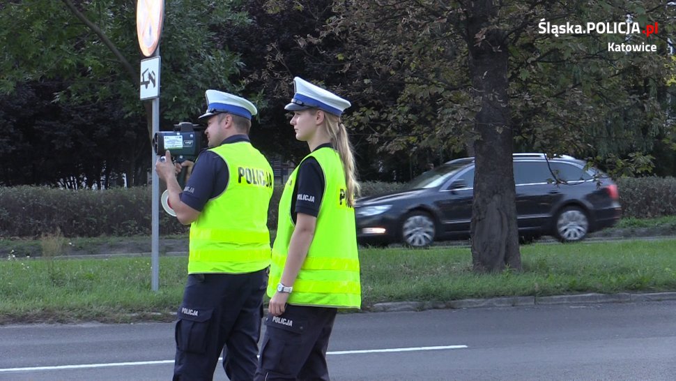 Na kolorowym zdjęciu widać policjanta i polciajntke w kamizelakch odbalskowych, któzy stoją przy ulicy i dokonują pomiaru prędkości