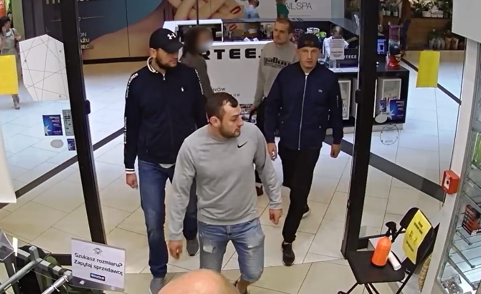 zdjęcie kolorowe: 4 mężczyzn podejrzewanych o kradzież broni i amunicji wchodzących do sklepu : pierwszy ubrany w jasnoniebieskie  jeansowe spodenki za kolano i szara bluzę dresowa z logo firmy Nike, drugi z mężczyzn ubrany w szare sportowe buty z biała podeszwą, granatowe długie spodnie jeansowe, granatową bluzę sportową z długim rękawem zapinaną na biały zamkiem i napisem na ramionach Ralph Lauren, granatowa czapkę z daszkiem z logo firmy Under Armour, trzeci z mężczyzn ubrany w czarne spodnie jeansowe, granatowa bluzę dresową zapinana na srebrny zamek i granatową czapkę z daszkiem założona na głowę daszkiem do tyłu, czwarty z mężczyzn ubrany w szara bluzę dresową z czarnym napisem.