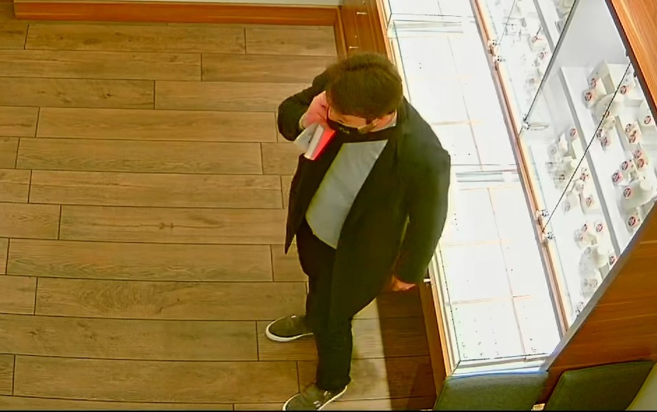zdjęcie kolorowe: screen z monitoringu sklepowego przedstawiający mężczyznę ubranego w ciemne spodnie, ciemny płaszcz, szary sweter z założona czarna maseczka ochronna na twarzy i rozmawiający przez telefon komórkowy, lewy profil