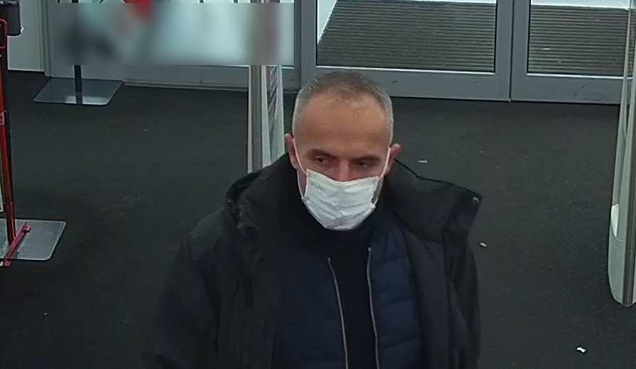 zdjęcie kolorowe: mężczyzna w średnim wieku ubrany w granatowe jeansy, czarna kurtkę z maseczką ochronna na twarzy wchodzący do marketu