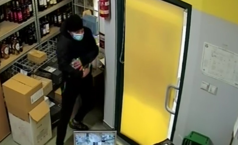 zdjęcie kolorowe: mężczyzna ubrany w czarne spodnie, czarna kurtkę i czarną czapkę z maseczka ochronną na twarzy w pokoju socjalnym sklepu spożywczego 