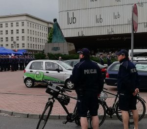 policjanci stoją przy rowerach przed nimi stoją samochody a za samochodami na placu stoją pododdziały Wojska i Policji biorące udział w uroczystości Święta Konstytucji 3 maja.