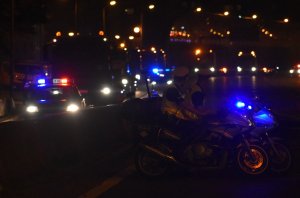 Na zdjęciu widać dwóch policjantów na motocyklach blokujących przejazd w tle jedzie wojskowa kolumna