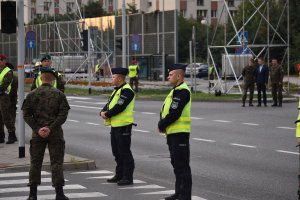 Na zdjęciu widać dwóch policjantów oraz żołnierzy jak stoją przy skrzyżowaniu w tle widać ministra obrony narodowej z generałami
