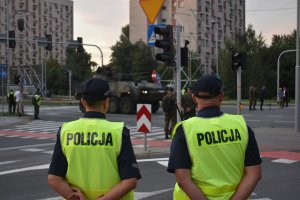 Na zdjęciu widać policjanta i policjantkę przed nimi jedzie transporter opancerzony w tle widać bloki przy uniwersyteckiej