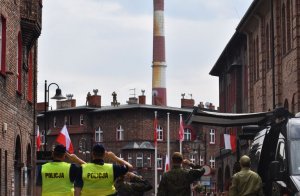 Na zdjęciu widać dwóch policjantów przednimi stoją ludzie w tel widać budynki i komin