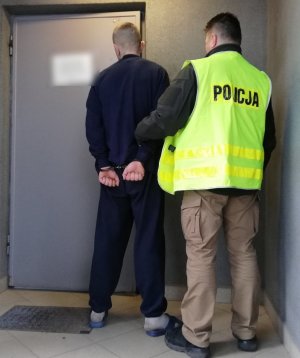 zdjęcie kolorowe: policjant kryminalny w kamizelce odblaskowej z napisem &quot;Policja&quot; z zatrzymanym mężczyzną podejrzanym o rozbój