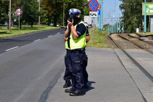 Na zdjęciu widać dwóch policjantów w kamizelkach odblaskowych, którzy stoją przy drodze i mierzą prędkość obok stoją znaki drogowe