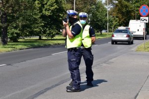 Na zdjęciu widać dwóch policjantów w kamizelkach odblaskowych, którzy stoją przy drodze i mierzą prędkość, za nimi jadą samochody