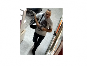 zdjęcie kolorowe: skan z monitoringu sklepowego przedstawiającego mężczyznę w wieku około 40 lat, posiadający krótkie siwiejące włosy z widocznymi zakolami, ubrany w szarą koszulkę z krótkimi rękawami posiadająca nadruk w kształcie czaszki, ciemne spodnie, czarne buty i czarno-biały plecak.