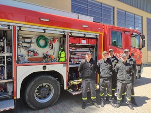 zdjęcie kolorowe: pięciu strażaków stojących przed wozem strażackim