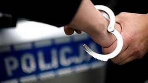zdjęcie kolorowe: na tle policyjnego radiowozu ręka, na którą zakładane są policyjne kajdanki