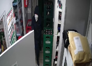 zdjęcie kolorowe: ubrany na czarno mężczyzna, w bluzie z kapturem  założonym na głowę, z zamaskowana twarzą chodzi po sklepie spożywczym