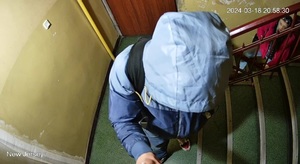zdjęcie kolorowe: mężczyzna stojący na klatce schodowej, ubrany w granatowe spodnie i niebieska kurtkę, z założonym kapturem na głowę