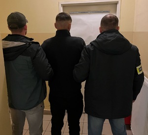 zdjęcie kolorowe: dwóch policjantów wydziału kryminalnego stoi na korytarzu z zatrzymanym mężczyzną